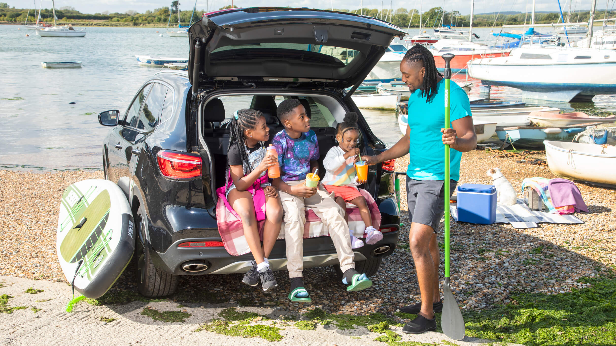 A family enjoys a summer day by the water, with three children sitting in the open boot of a black SUV drinking juice. An adult stands beside them holding a paddle, with paddleboards and boats visible in the background. A dog relaxes on a blanket nearby, surrounded by coolers and towels on the pebbled shore.