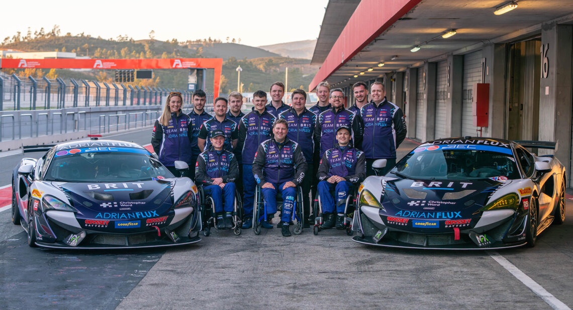 Team BRIT members pose for a group photo in front of two racing cars in a pit lane. Several team members use wheelchairs, and all are wearing matching navy racing uniforms.