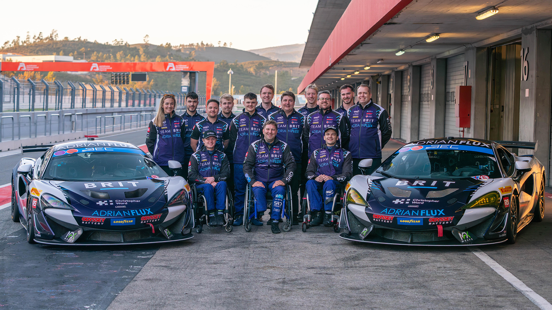 Team BRIT members pose for a group photo in front of two racing cars in a pit lane. Several team members use wheelchairs, and all are wearing matching navy racing uniforms.