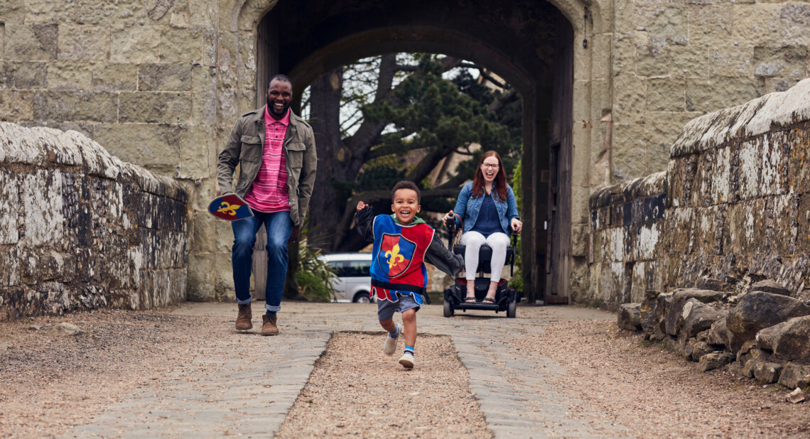 A young boy dressed as a knight runs joyfully across a stone bridge, followed by a smiling man holding toy shields and a woman in a powered wheelchair, with a castle archway behind them.