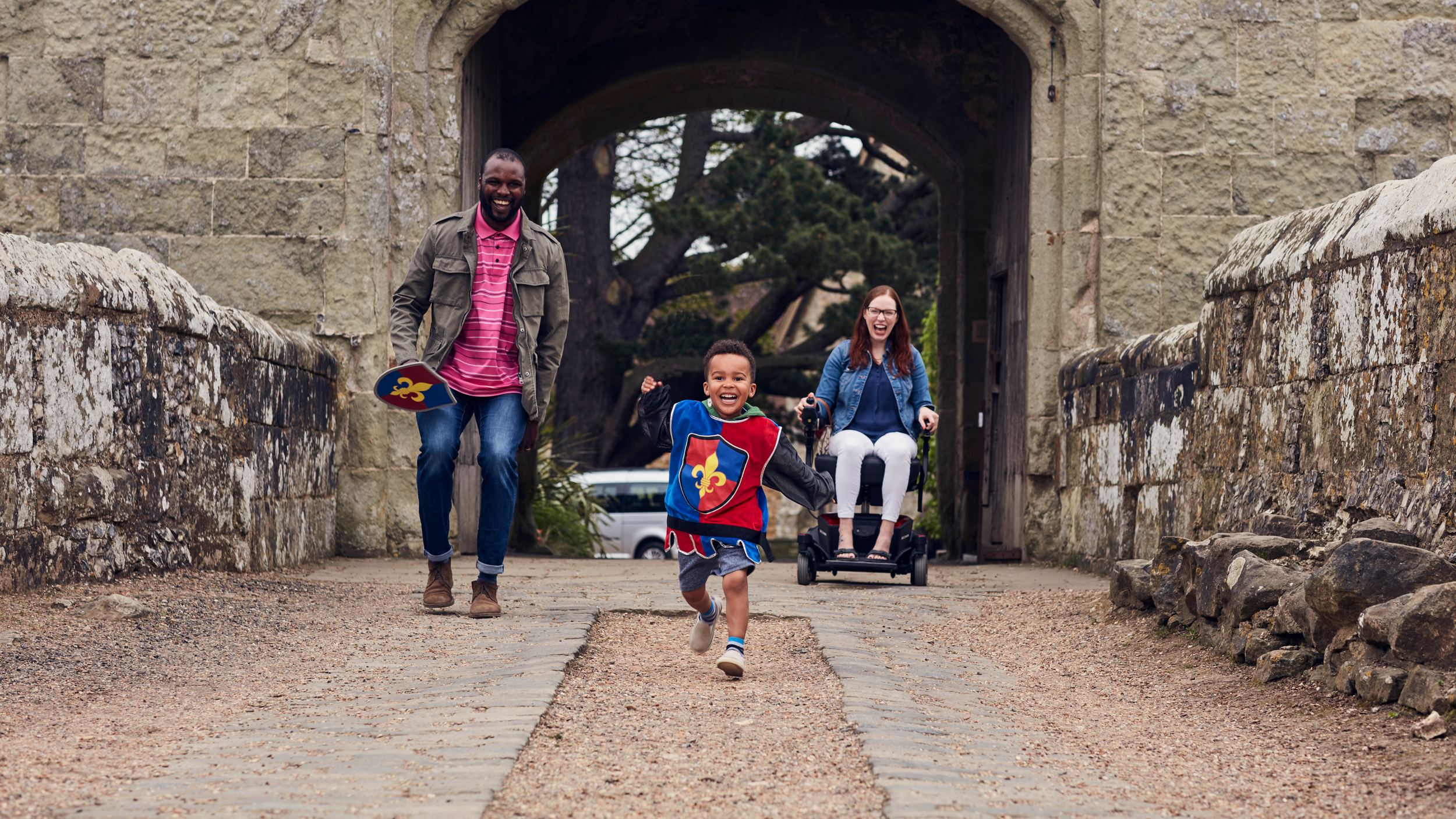 A young boy dressed as a knight runs joyfully across a stone bridge, followed by a smiling man holding toy shields and a woman in a powered wheelchair, with a castle archway behind them.