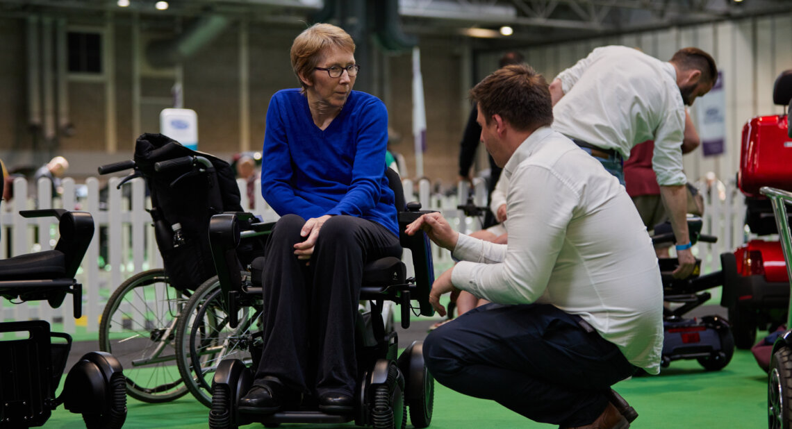 A man kneels to talk to a woman sitting in a powered wheelchair at Motability Scheme Live.