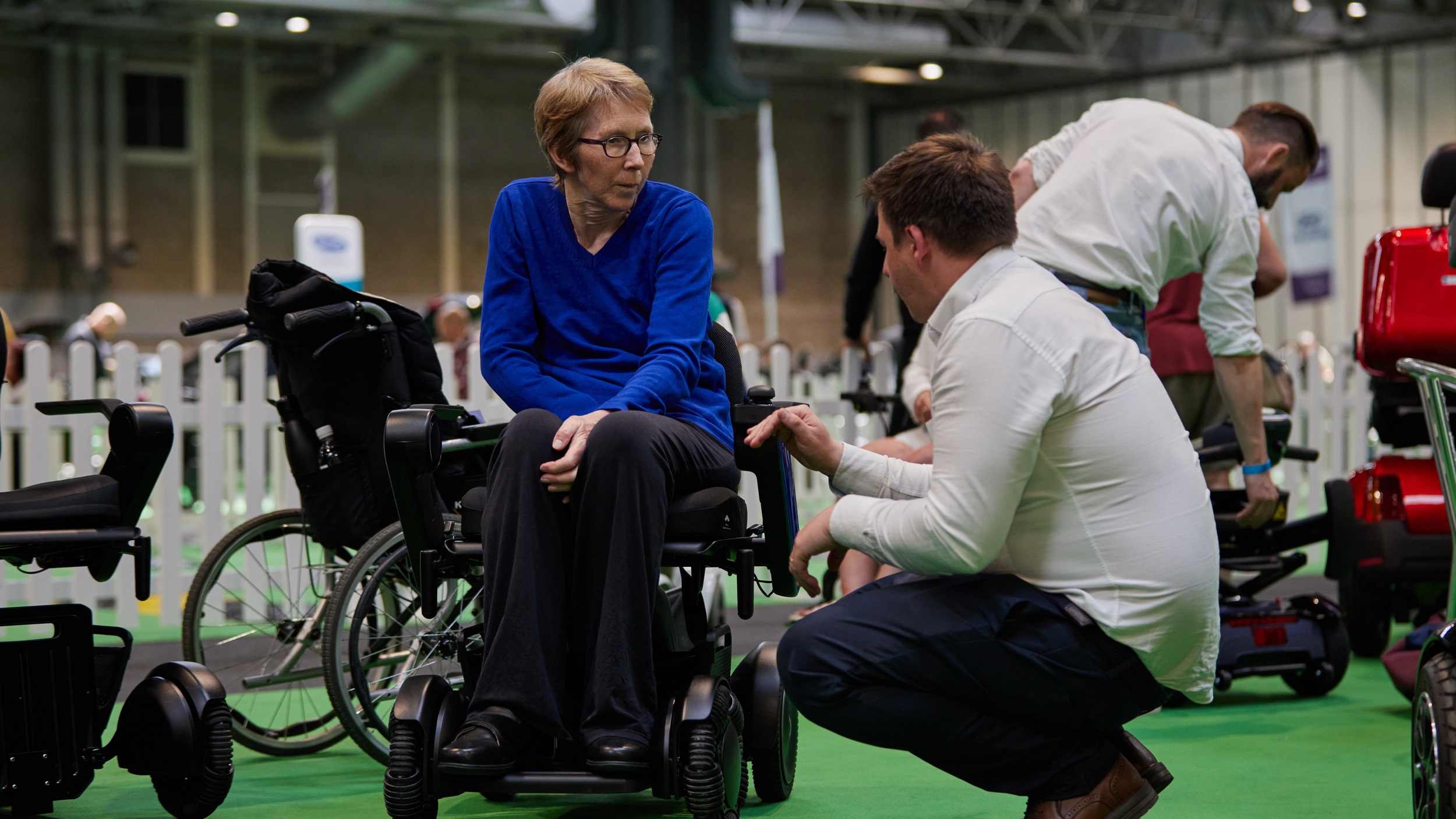 A man kneels to talk to a woman sitting in a powered wheelchair at Motability Scheme Live.