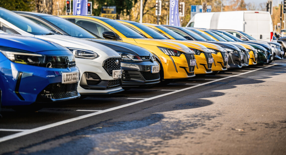 A row of parked cars in various colours including blue, white, grey, and yellow, neatly lined up in a dealership lot on a sunny day.
