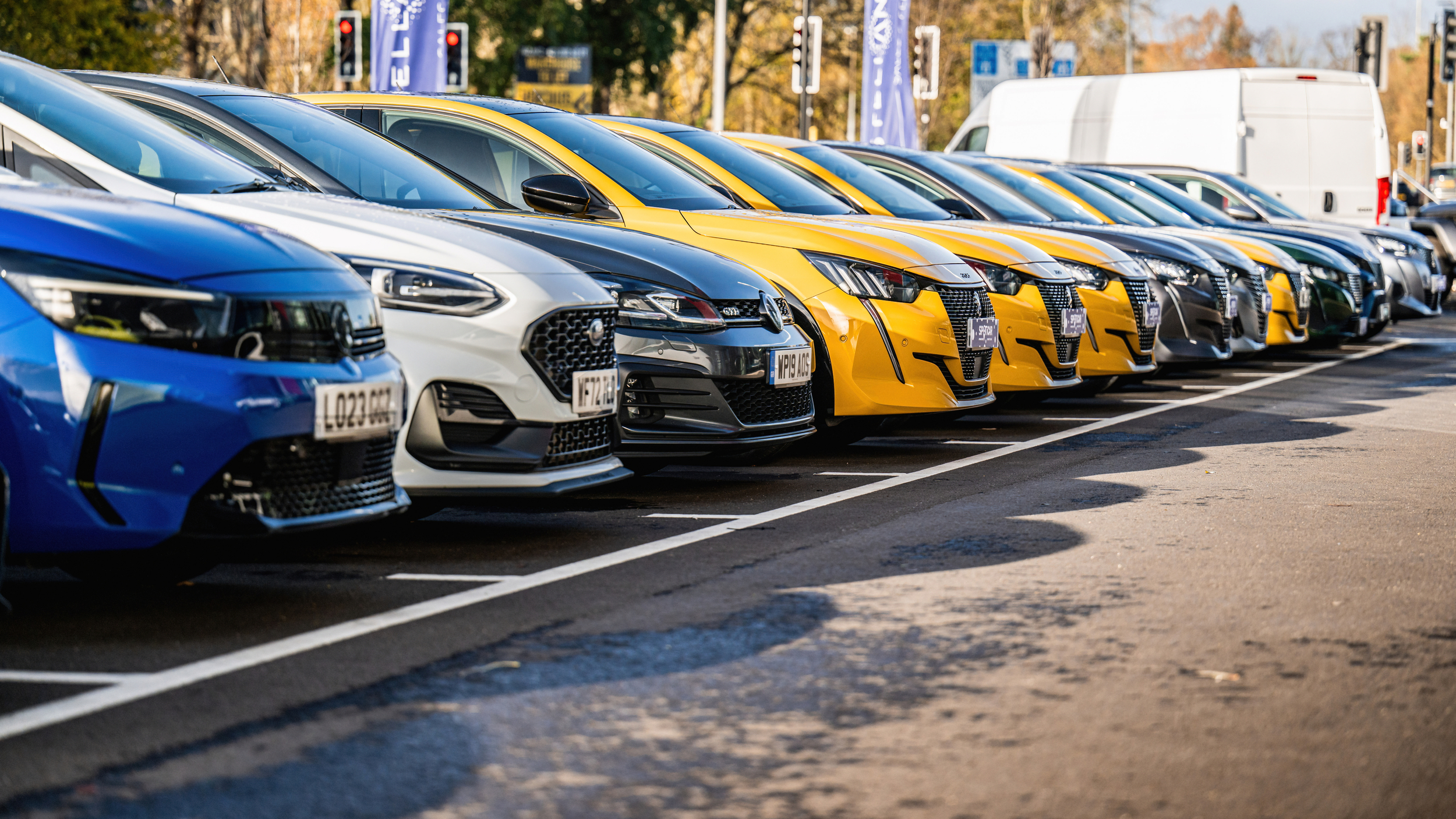 A row of parked cars in various colours including blue, white, grey, and yellow, neatly lined up in a dealership lot on a sunny day.
