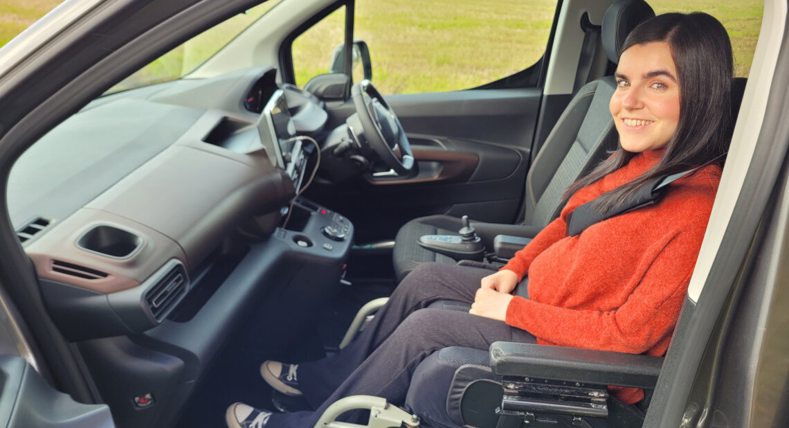 Emma Muldoon smiles while seated in the front passenger position of a WAV, secured in her powered wheelchair inside the adapted vehicle.