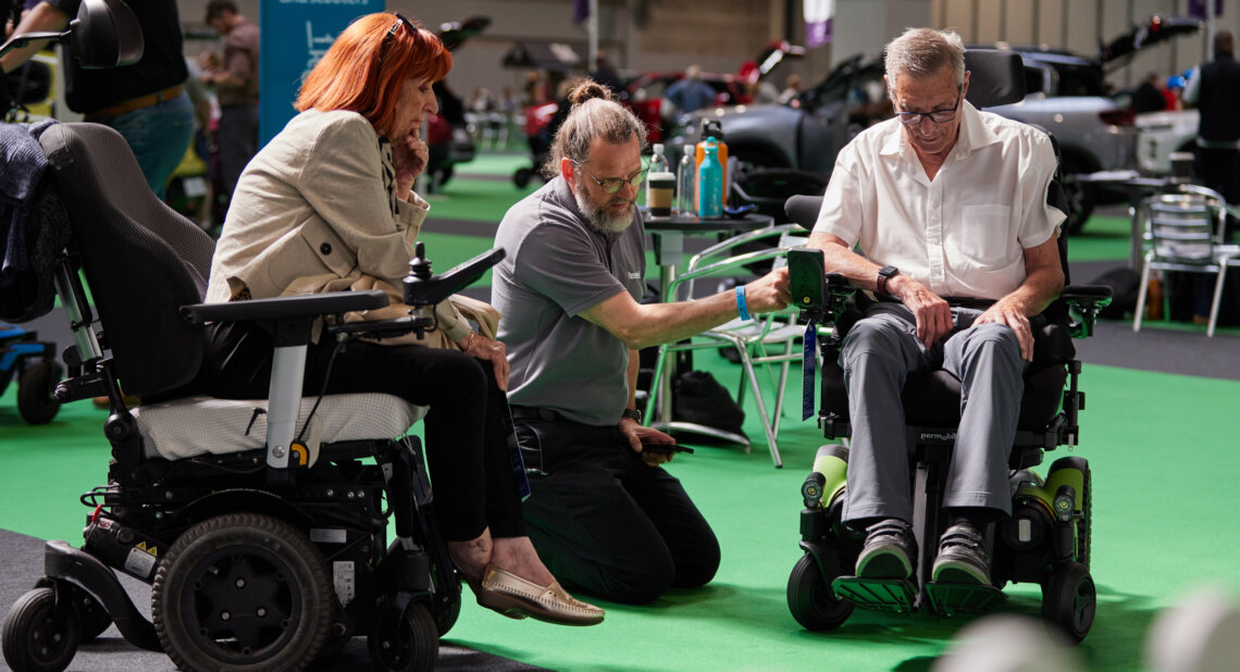 An exhibitor kneels between two older adults in powered wheelchairs at an indoor mobility event, demonstrating joystick controls. A sign in the background reads "Powered wheelchairs and scooters," and adapted vehicles are visible throughout the venue.
