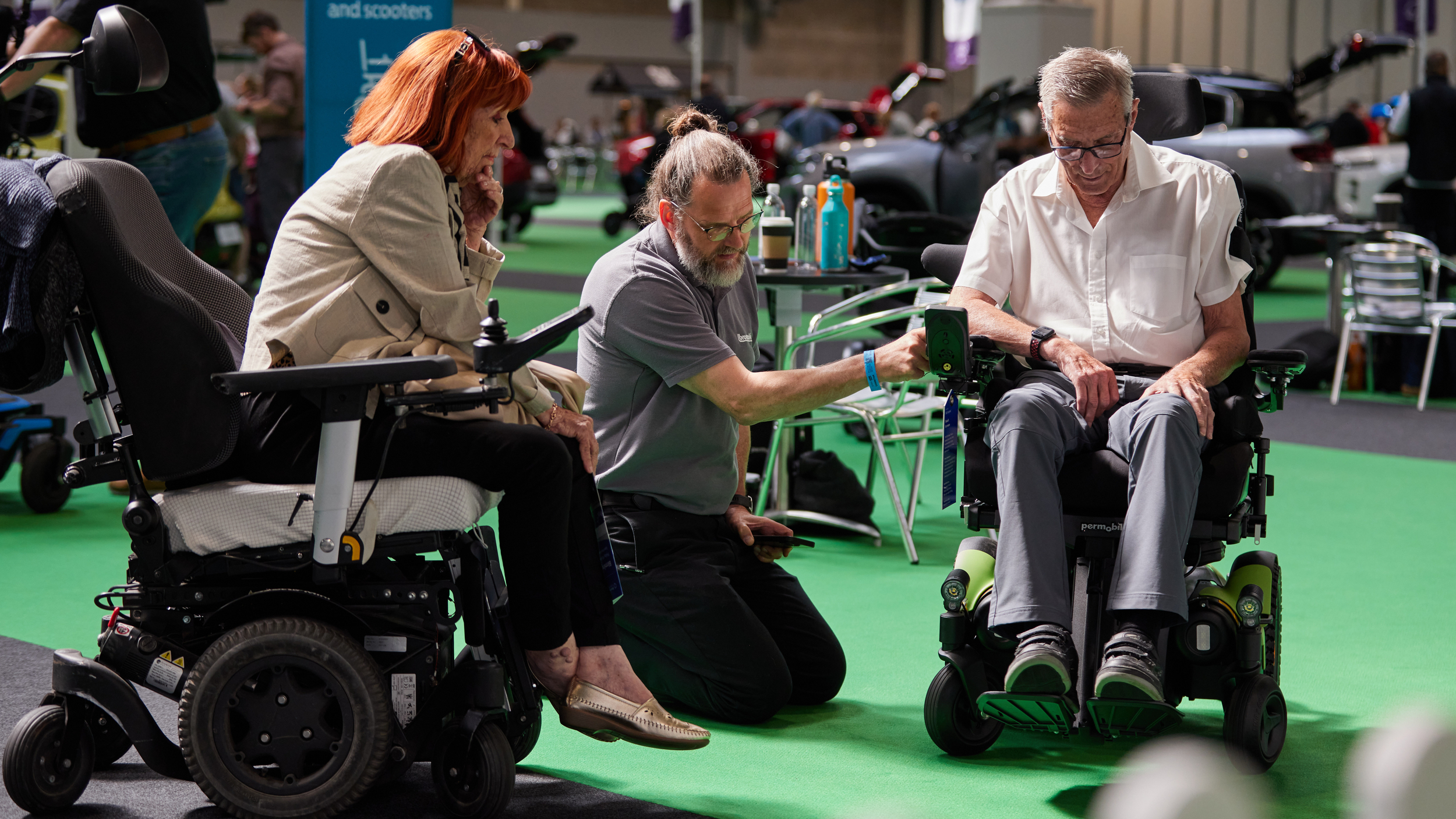 An exhibitor kneels between two older adults in powered wheelchairs at an indoor mobility event, demonstrating joystick controls. A sign in the background reads "Powered wheelchairs and scooters," and adapted vehicles are visible throughout the venue.