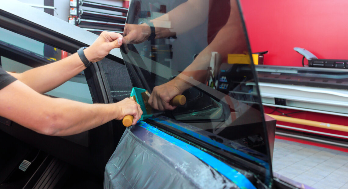 A person installs tinted privacy film on a car window using a squeegee tool, pressing the film smoothly onto the glass inside a workshop.