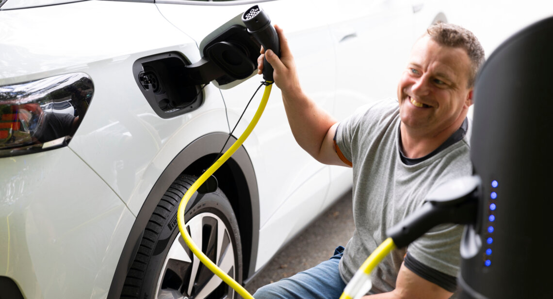 A smiling man kneels beside a white electric vehicle as he prepares to plug in a yellow charging cable at a public charging station.