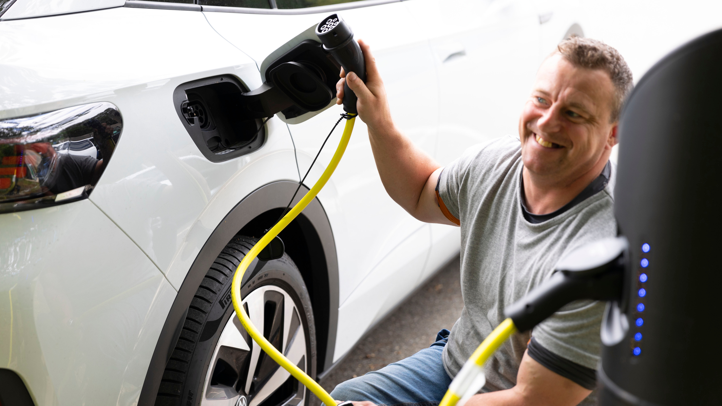 A smiling man kneels beside a white electric vehicle as he prepares to plug in a yellow charging cable at a public charging station.