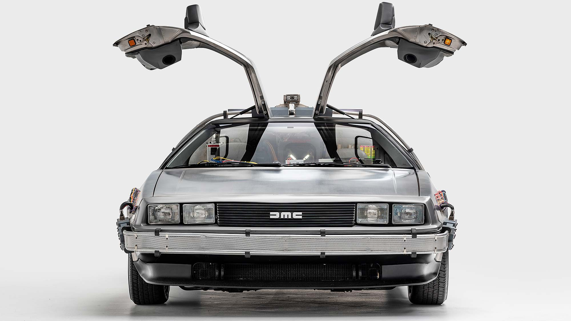 A modified DeLorean DMC-12 from the Back to the Future film, shown head-on with gullwing doors open and time-travel electronics visible inside and on the bumper.