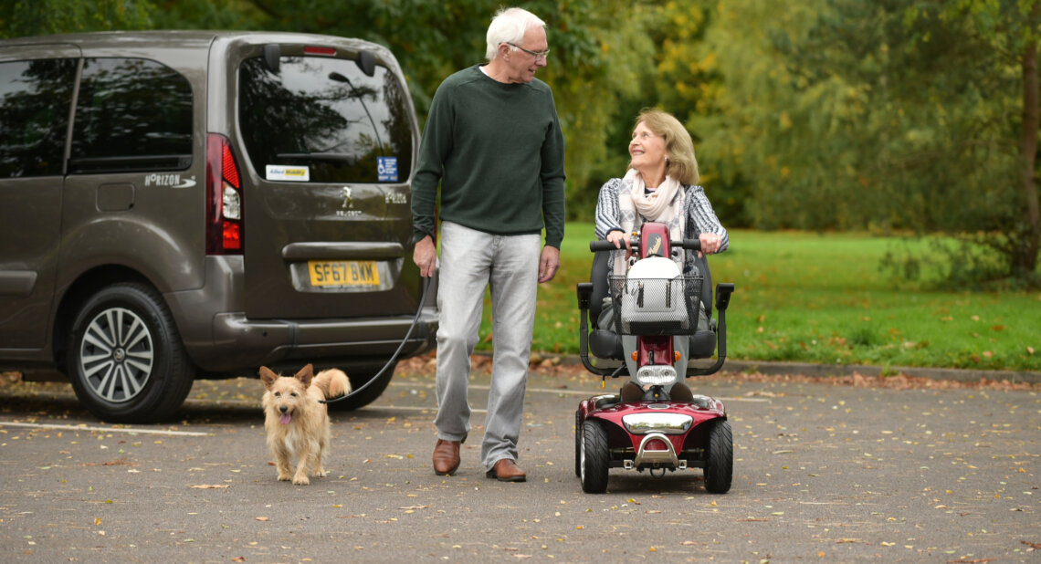 An older man walks a small dog beside a woman using a red mobility scooter in a leafy car park. A wheelchair accessible vehicle (WAV) with a Blue Badge and Allied Mobility stickers is parked nearby.