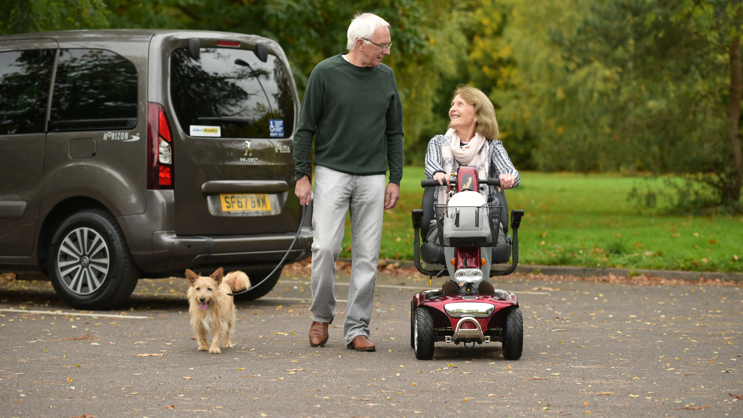 An older man walks a small dog beside a woman using a red mobility scooter in a leafy car park. A wheelchair accessible vehicle (WAV) with a Blue Badge and Allied Mobility stickers is parked nearby.