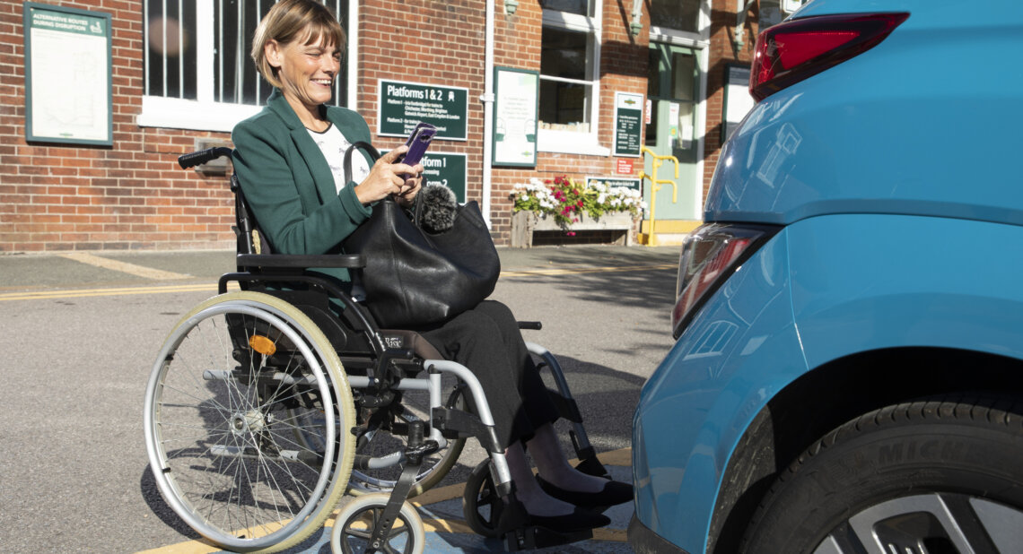 A woman in a manual wheelchair smiles while using her smartphone in a car park outside a train station, positioned near the rear of a blue car parked in a Blue Badge bay.