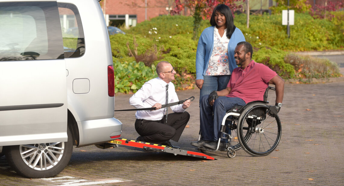 A man in a wheelchair smiles as a mobility advisor demonstrates the rear ramp of a silver wheelchair accessible vehicle (WAV), while a woman stands nearby, watching the demonstration in a sunny car park.