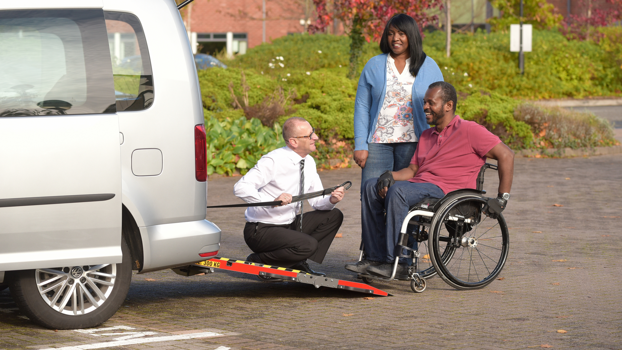 A man in a wheelchair smiles as a mobility advisor demonstrates the rear ramp of a silver wheelchair accessible vehicle (WAV), while a woman stands nearby, watching the demonstration in a sunny car park.