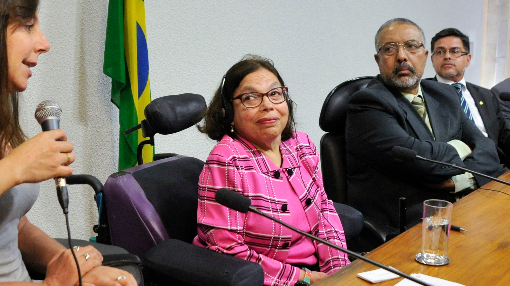 Disability rights activist Judith Heumann sits in a power wheelchair at a formal panel discussion, wearing a pink plaid jacket and headset, alongside three suited men and a Brazilian flag in the background.
