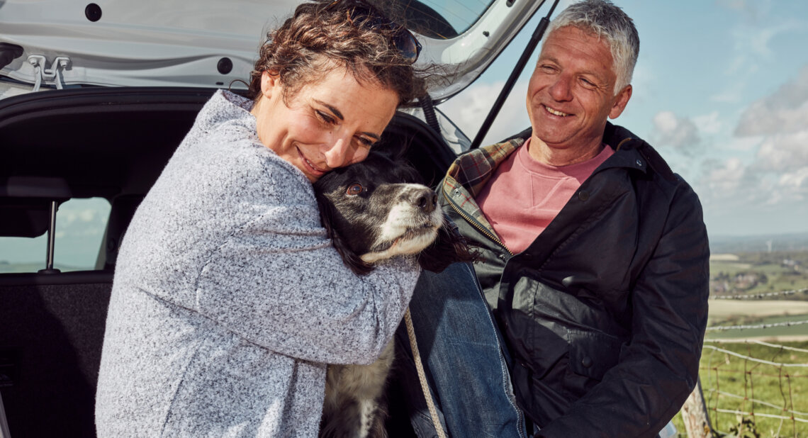 A woman hugs a black and white spaniel at the open boot of a car while a man smiles beside her. They are outdoors in a countryside setting on a bright, breezy day.