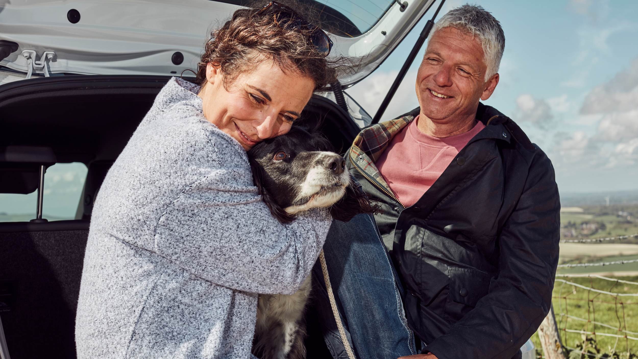 A woman hugs a black and white spaniel at the open boot of a car while a man smiles beside her. They are outdoors in a countryside setting on a bright, breezy day.
