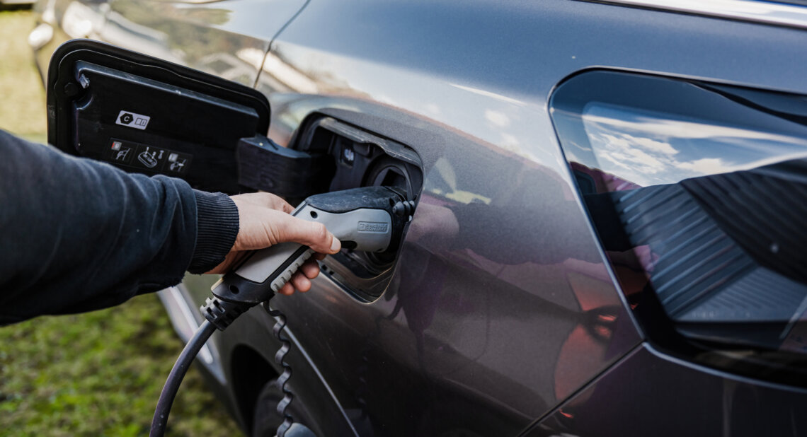 A close-up of a person’s hand plugging an electric vehicle (EV) charging cable into the charging port of a dark grey car, with grass and sky reflected in the car's surface.