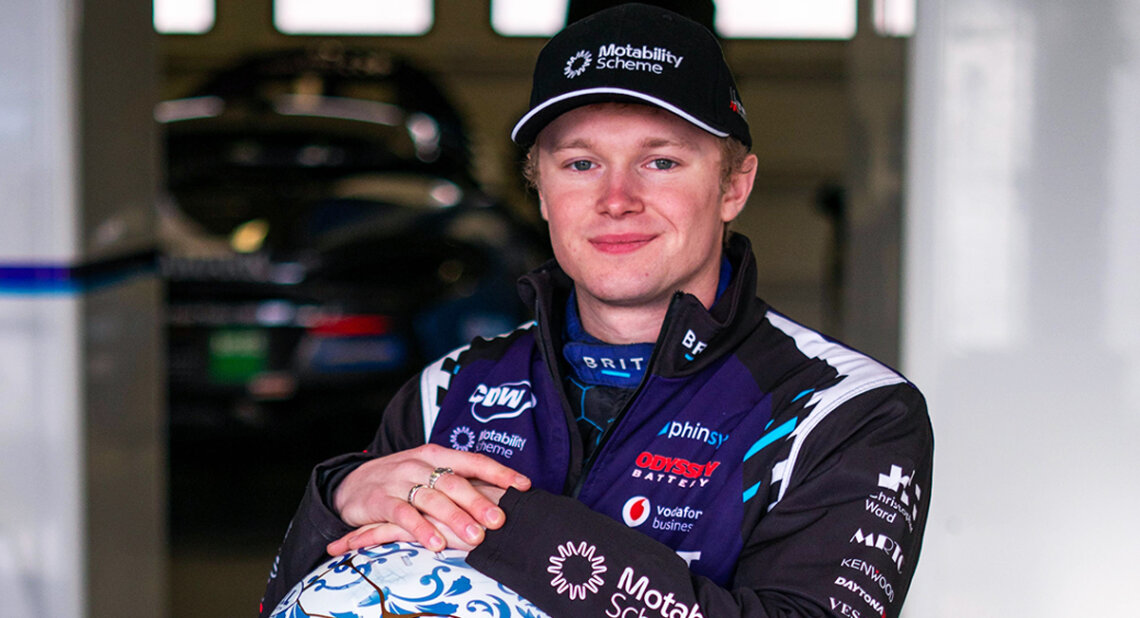 Noah Cosby in racing gear and a Motability Scheme cap poses with a patterned racing helmet, smiling confidently in a garage with blurred race cars in the background.