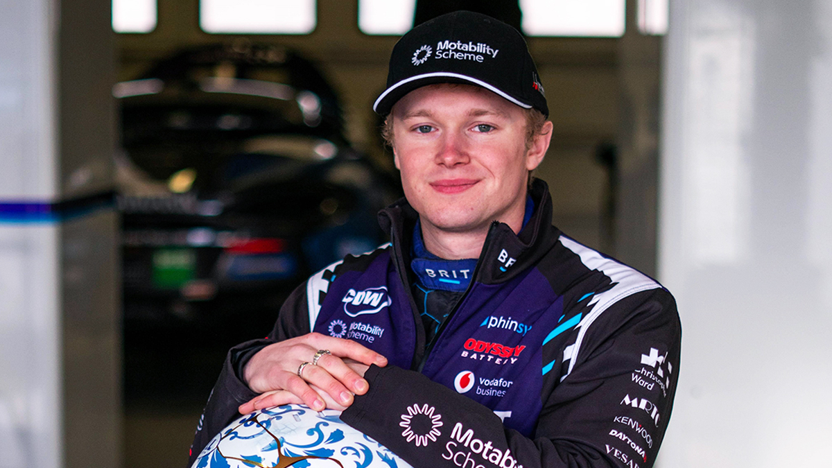 Noah Cosby in racing gear and a Motability Scheme cap poses with a patterned racing helmet, smiling confidently in a garage with blurred race cars in the background.