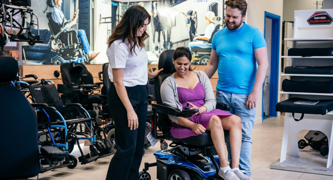 A smiling woman tests a powered wheelchair in a mobility showroom, assisted by a staff member and a man standing beside her. Various mobility aids are displayed around them.