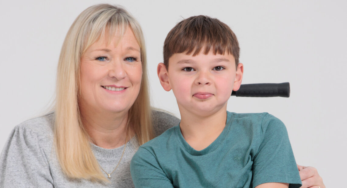 Paula Hudgell, with long blonde hair and a grey top, smiles warmly beside her son Tony, who wears a teal T-shirt and sits in his wheelchair. Tony looks at the camera with a playful expression, his tongue slightly out.