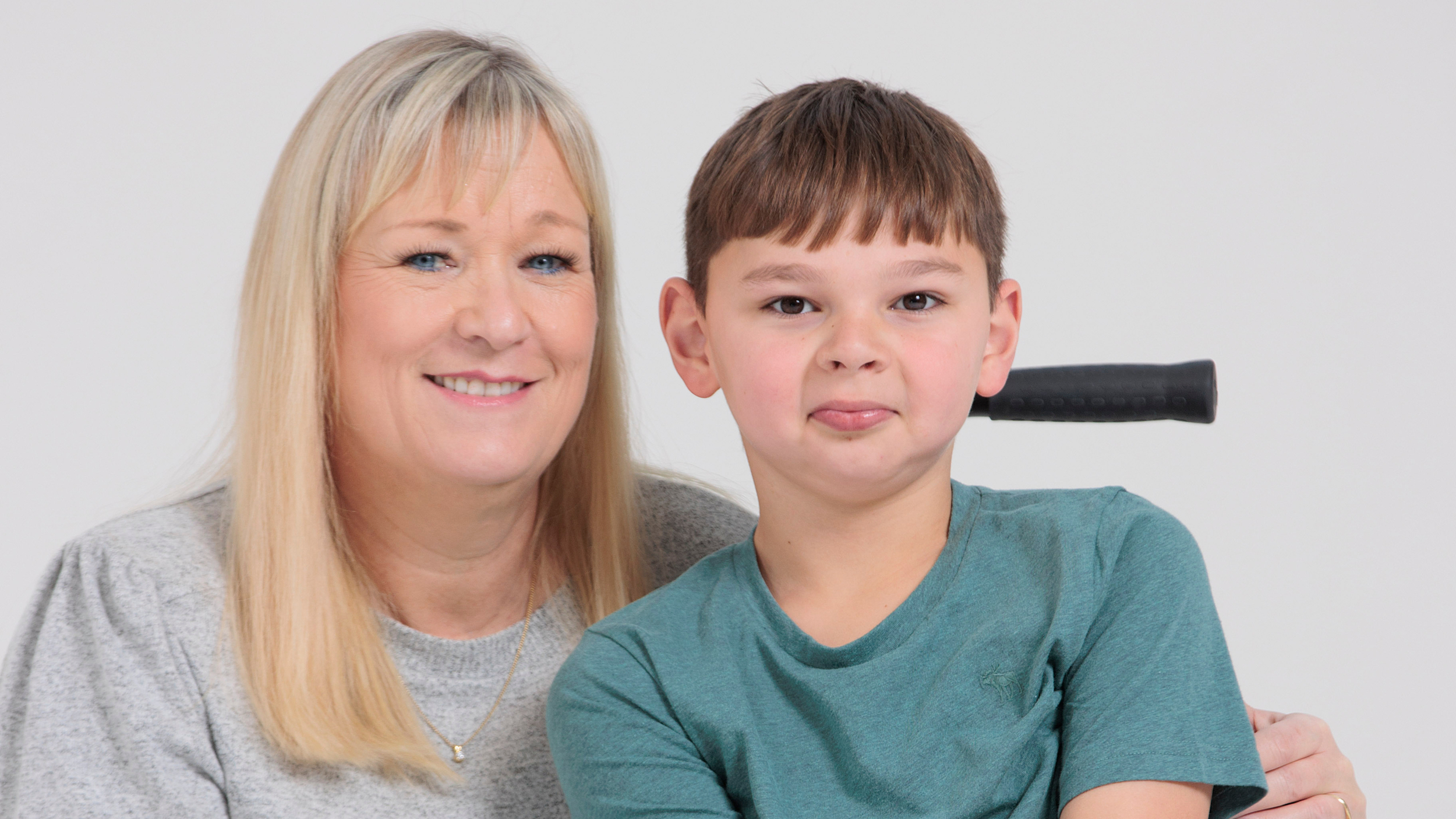 Paula Hudgell, with long blonde hair and a grey top, smiles warmly beside her son Tony, who wears a teal T-shirt and sits in his wheelchair. Tony looks at the camera with a playful expression, his tongue slightly out.