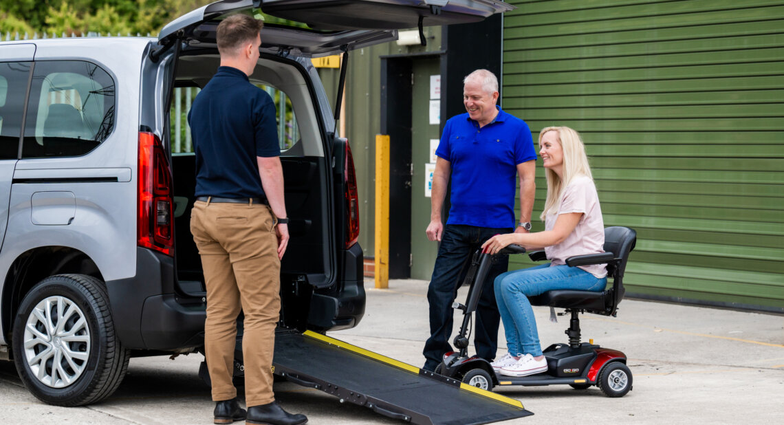 A woman on a red mobility scooter smiles as she approaches the rear ramp of a wheelchair accessible vehicle (WAV), while a man in a blue shirt and a mobility advisor stand nearby offering support.