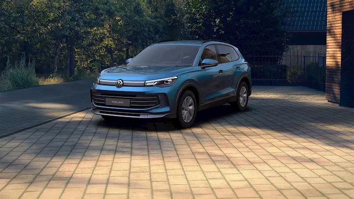 A blue Volkswagen Tiguan SUV parked on a paved driveway surrounded by trees and a modern home, in soft natural lighting.