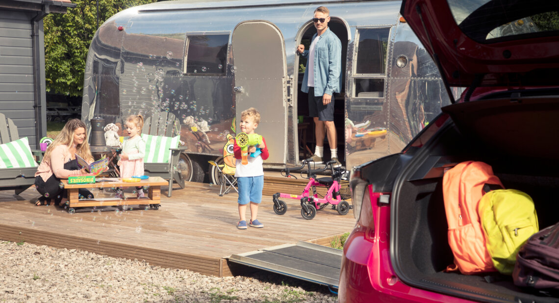 A family enjoying time outside a shiny silver caravan. A woman and young girl play at a small table, while a boy with a bubble machine stands nearby. A man watches from the caravan doorway. In the foreground, the open boot of a red car shows brightly coloured backpacks.