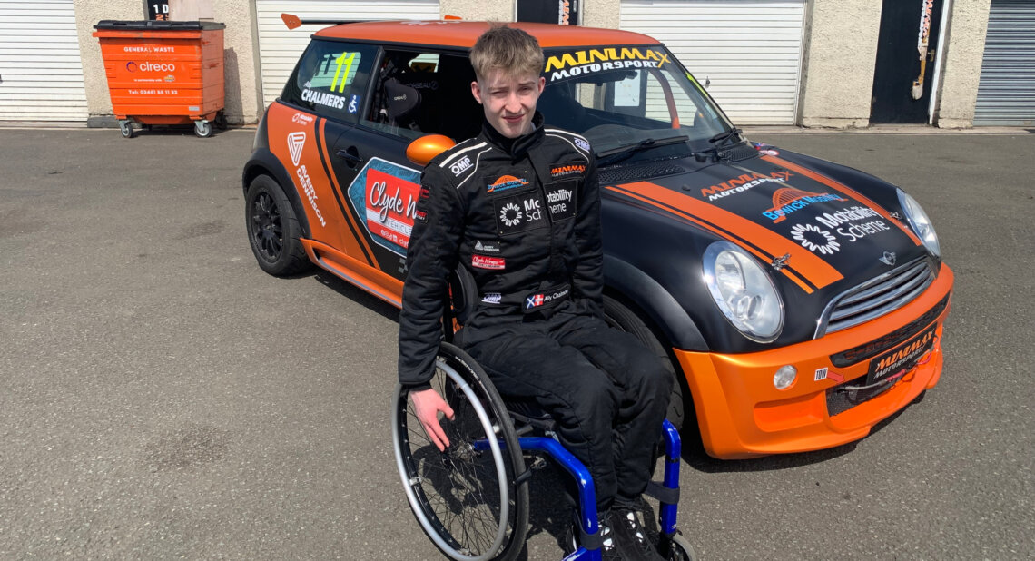 Ally Chalmers, a young male racing driver in a wheelchair, wearing a black racing suit with sponsor logos including the Motability Scheme, sits in front of his black and orange Mini Cooper race car, which is parked on a tarmac surface near garage doors