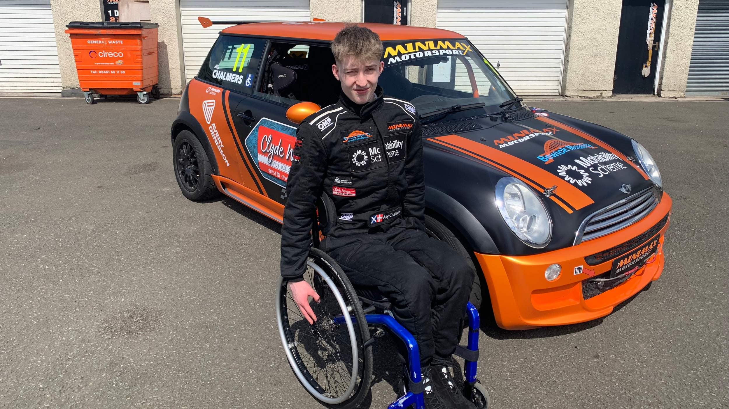 Ally Chalmers, a young male racing driver in a wheelchair, wearing a black racing suit with sponsor logos including the Motability Scheme, sits in front of his black and orange Mini Cooper race car, which is parked on a tarmac surface near garage doors
