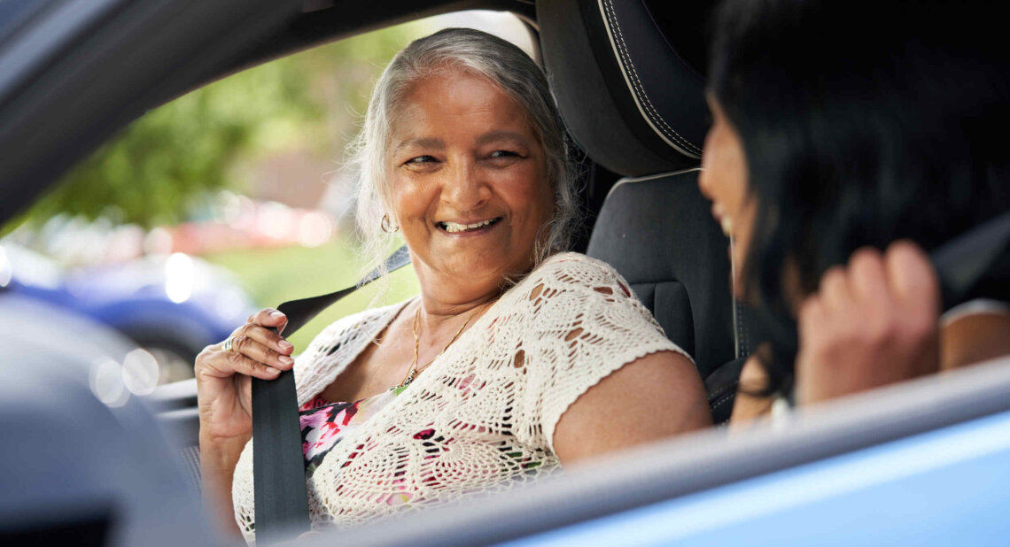 An older woman with long grey hair smiles warmly at another person while fastening her seatbelt inside a car. The two appear to be sharing a happy moment together before a drive.
