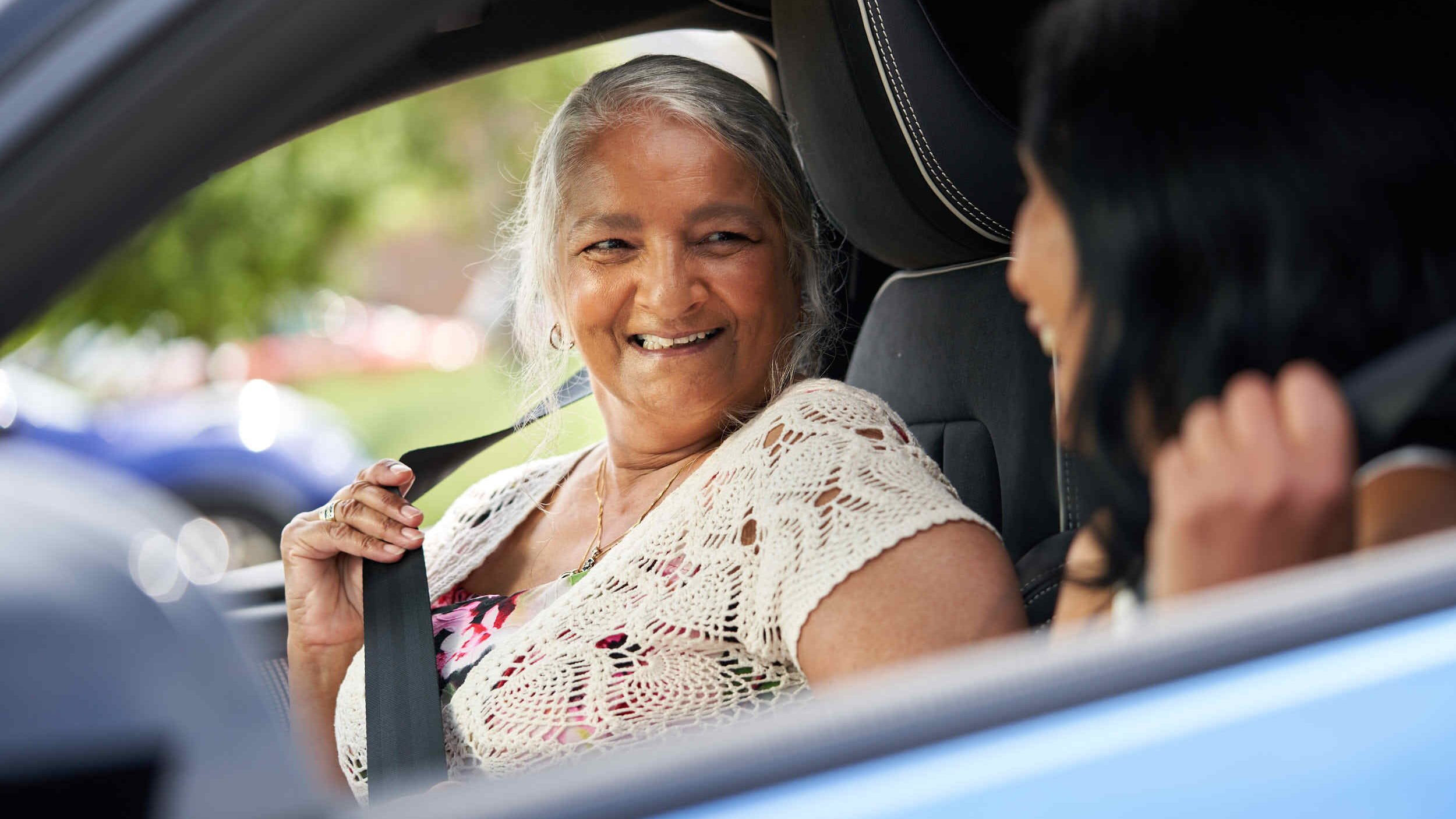 An older woman with long grey hair smiles warmly at another person while fastening her seatbelt inside a car. The two appear to be sharing a happy moment together before a drive.