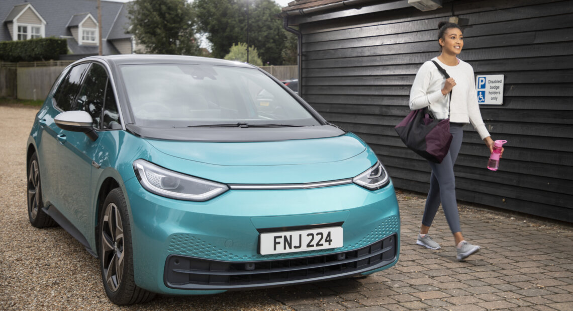 A turquoise electric car parked in front of a wooden building, with a woman carrying a gym bag and water bottle walking past.