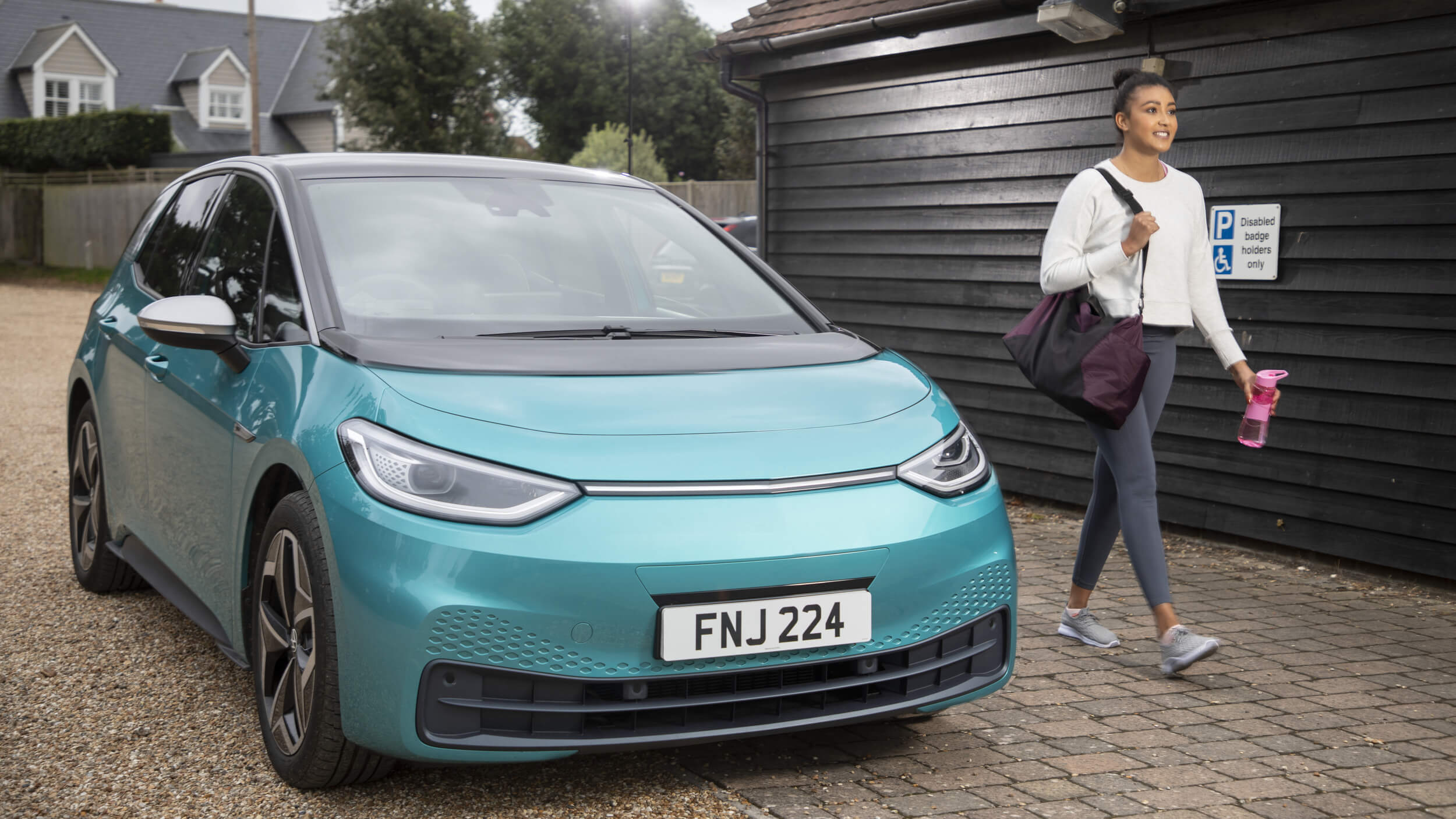 A turquoise electric car parked in front of a wooden building, with a woman carrying a gym bag and water bottle walking past.