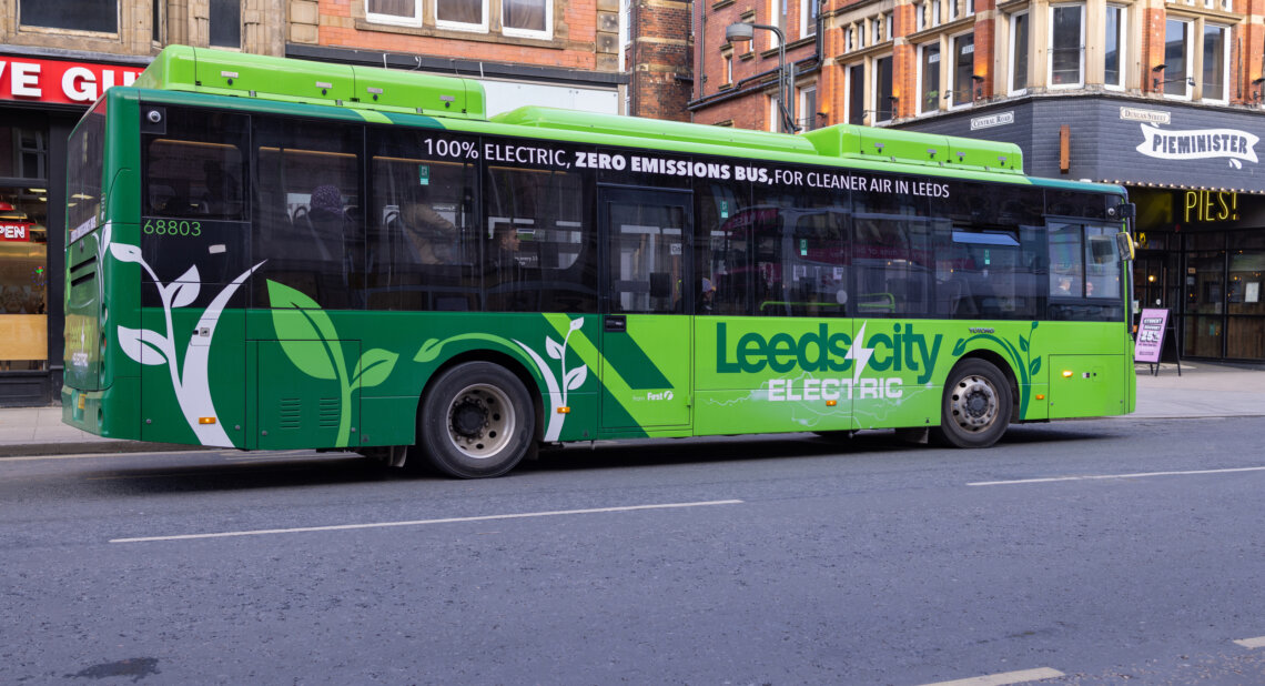 A bright green electric bus with leaf graphics and the words “Leeds City Electric” on the side is parked on a city street, promoting zero-emission transport in Leeds.