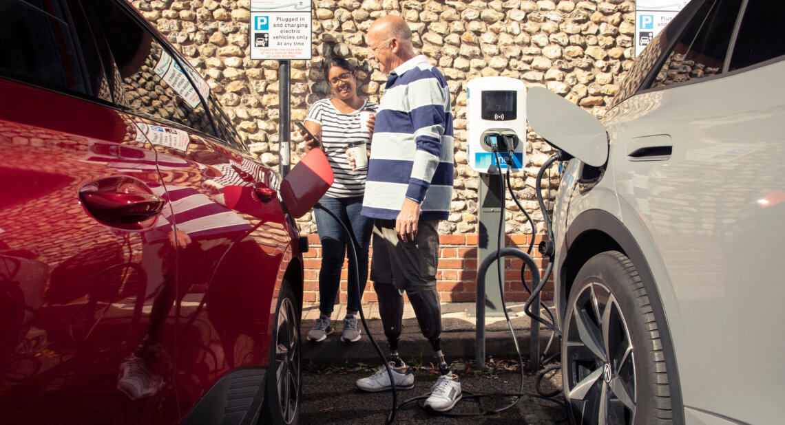 A man with prosthetic legs plugs an electric car into a charging point while a woman beside him smiles and holds a phone.