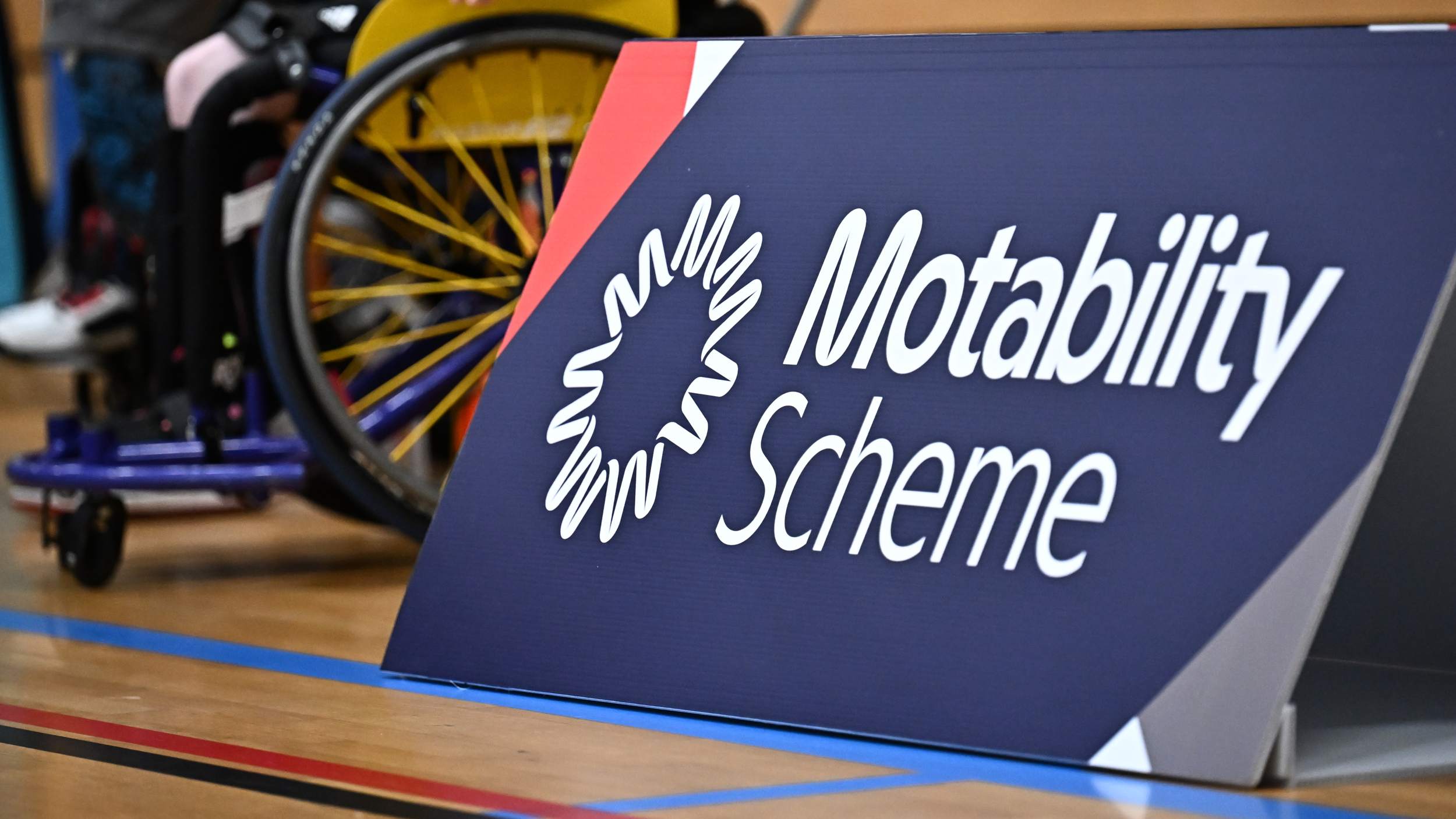 Close-up of a Motability Scheme courtside sign, with a wheelchair and colourful wheel spokes visible in the background.