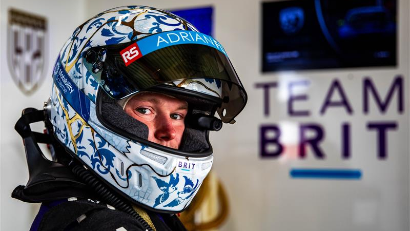 Close-up of racing driver Noah Cosby wearing a decorated helmet with a visor labelled “Adrian Flux,” standing in front of a wall with the words “Team Brit.