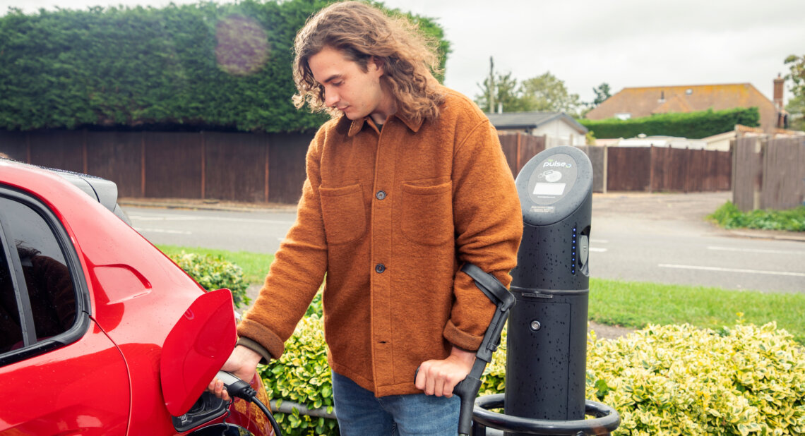 A person in a brown jacket charges a red electric car at a public charging point on a cloudy day.