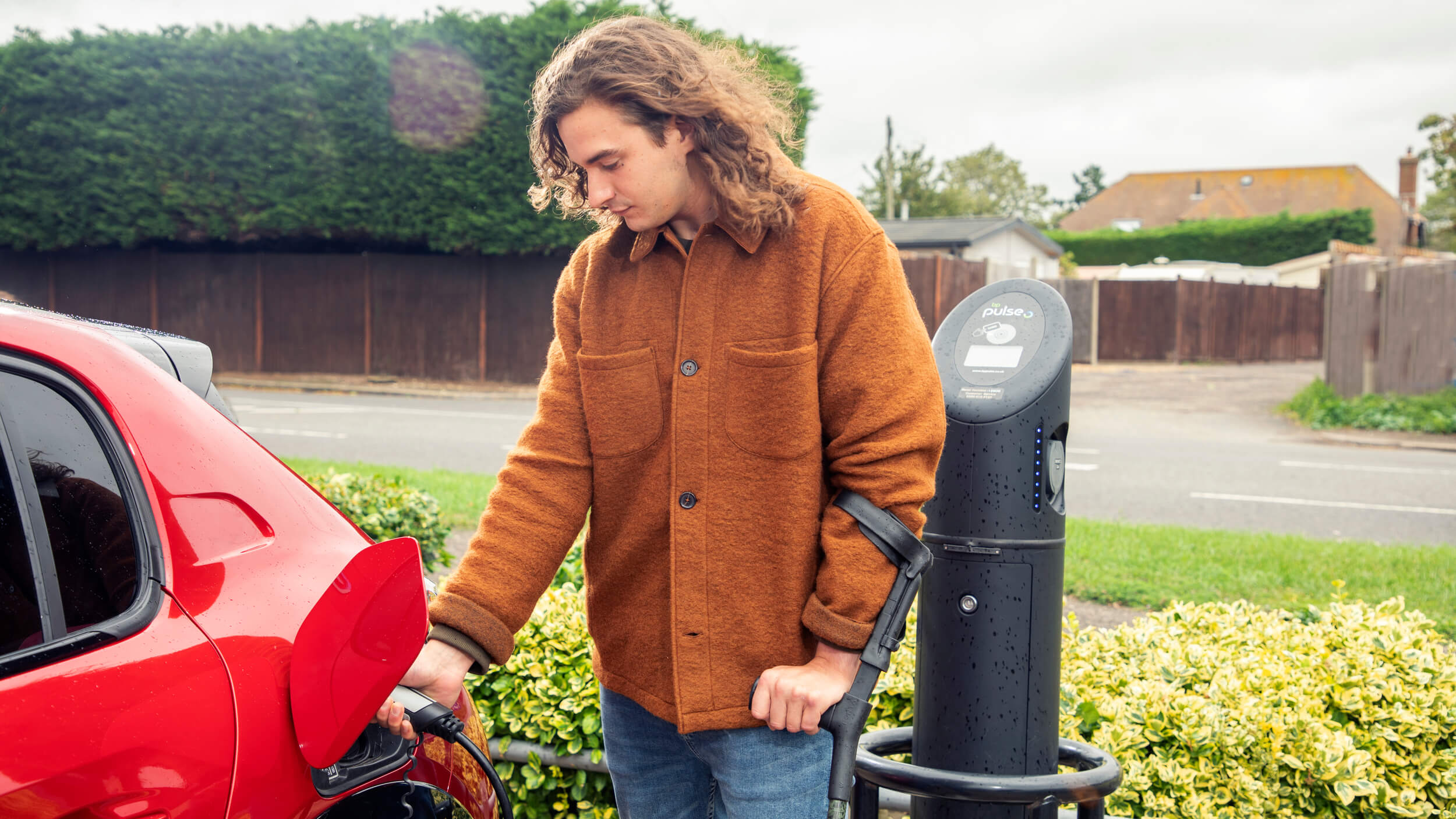 A person in a brown jacket charges a red electric car at a public charging point on a cloudy day.