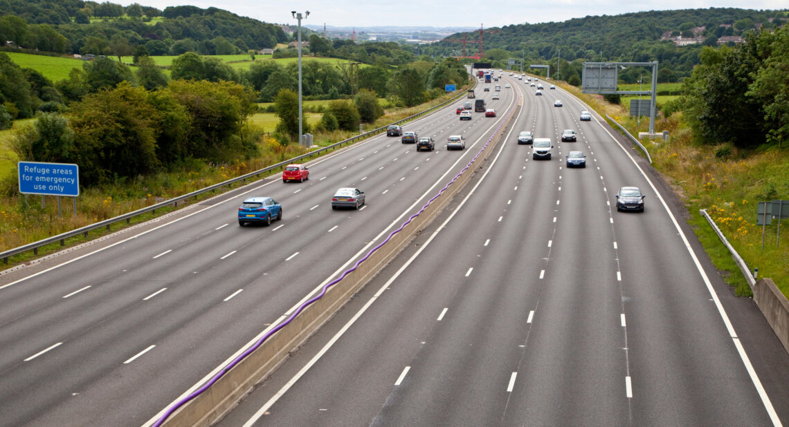 The M1 motorway with light traffic moving in both directions, surrounded by green fields and hills. A blue roadside sign reads “Refuge areas for emergency use only.”