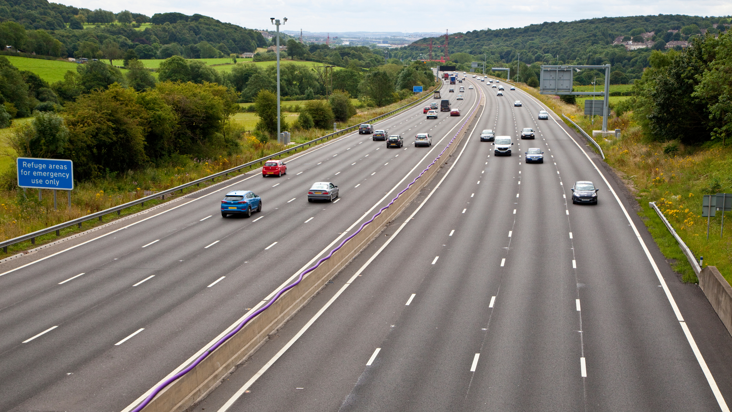 The M1 motorway with light traffic moving in both directions, surrounded by green fields and hills. A blue roadside sign reads “Refuge areas for emergency use only.”