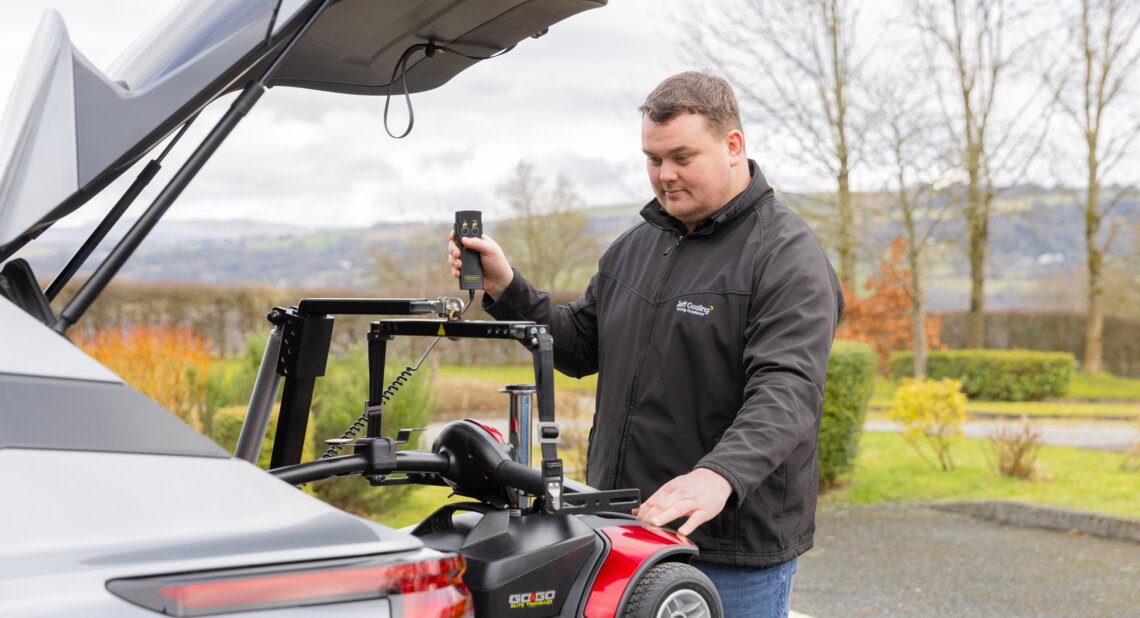 A man operating a Jeff Gosling electric hoist to lift a red mobility scooter into the boot of a grey SUV. He stands beside the open boot, holding the hoist’s remote control, with trees and countryside in the background.