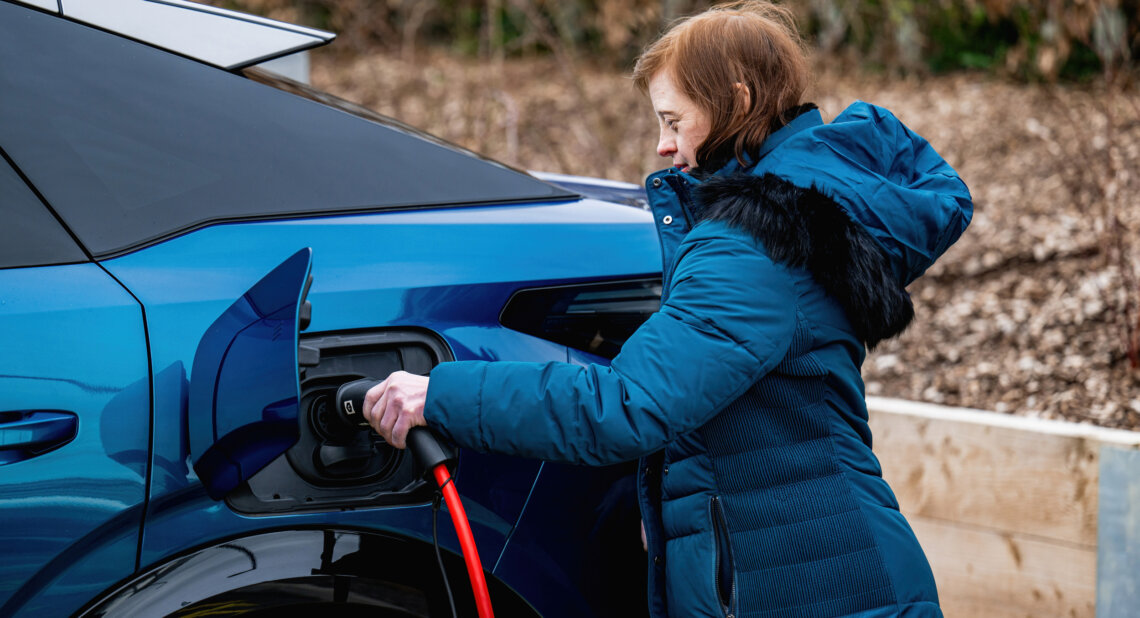 A woman wearing a blue winter coat plugs a charging cable into a blue electric car parked outdoors.