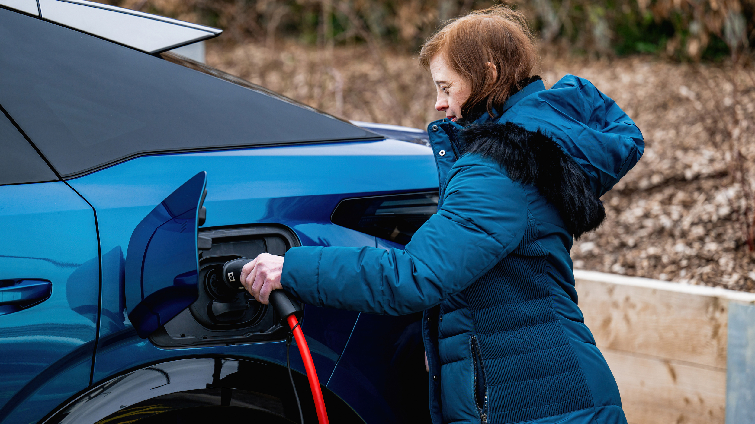 A woman wearing a blue winter coat plugs a charging cable into a blue electric car parked outdoors.